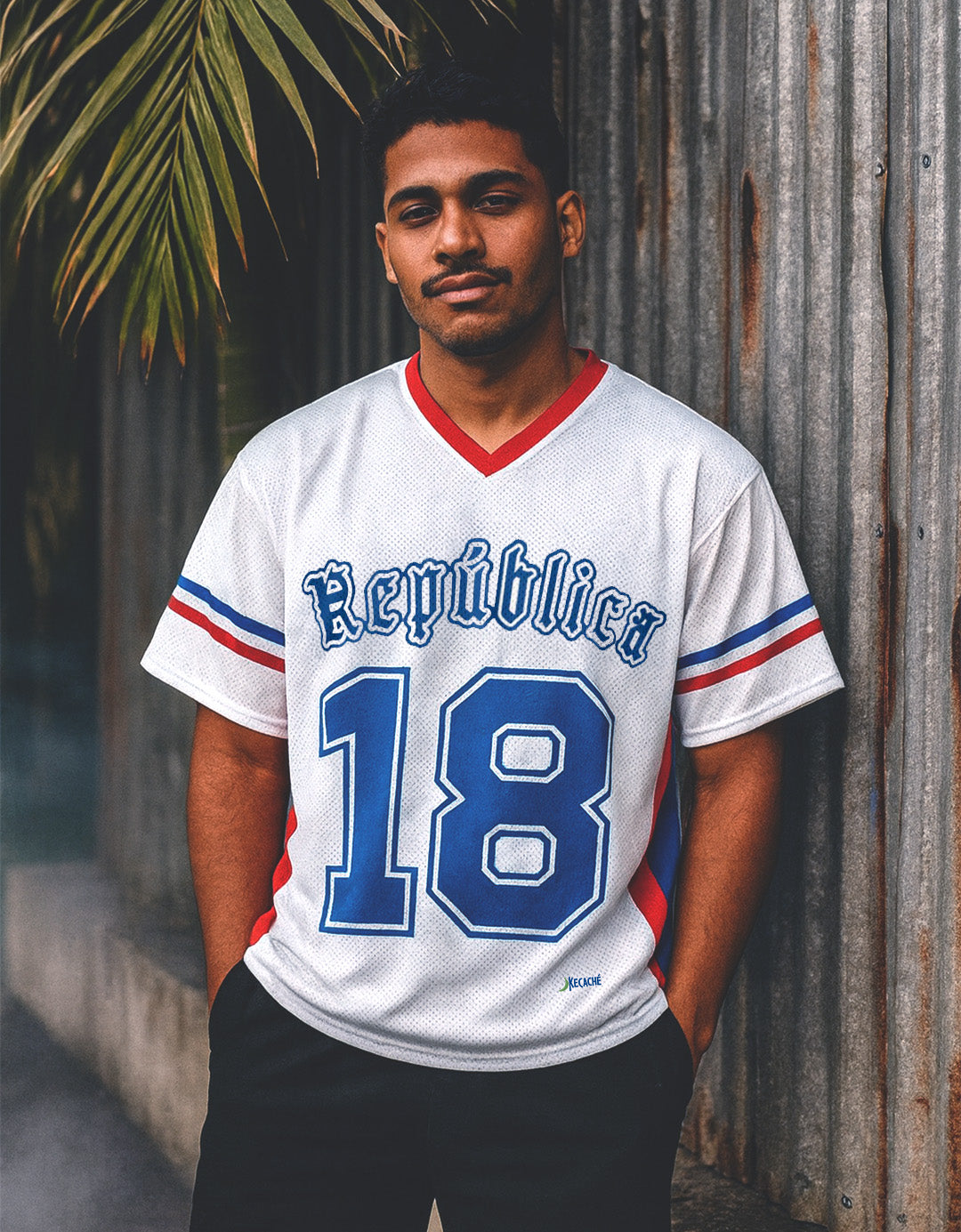 Man wearing a white sports Dominican jersey with blue and red details, standing against a rustic wall.
