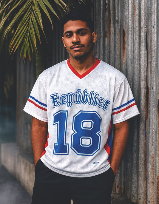 Man wearing a white sports Dominican jersey with blue and red details, standing against a rustic wall.